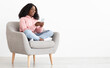 © Prostock-studio - Cheerful african american young lady sitting in arm chair at home, using nice mobile application on brand new smartphone, shopping online, white background, panorama with copy space