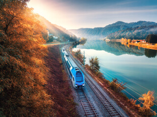 Aerial view of blue modern high speed train moving near lake in alpine mountains at sunrise in autumn. Top view of train, railroad, water, reflection, orange trees in fall. Railway station in Slovenia