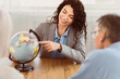 © Prostock-studio - Choosing Trip. Beautiful mexican agent proposing last minute travel offer to mature couple, pointing at globe, copyspace