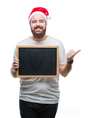 Wall Mural - Young caucasian hipster man wearing christmas hat holding blackboard over isolated background pointing and showing with thumb up to the side with happy face smiling