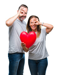 Canvas Print - Middle age hispanic casual couple in love holding red heart over isolated background with happy face smiling doing ok sign with hand on eye looking through fingers