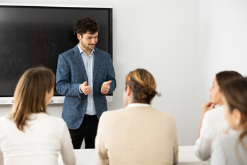 Wall Mural - Young male teacher near interactive board, gesturing with hand while consecrating topic of lesson for adults in classroom