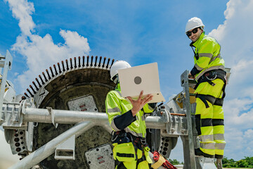 two engineers work on a wind turbine component, with one using a laptop and the other inspecting par