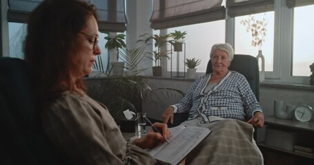 Sticker - In psychotherapist's office: Female specialist and elderly female patient sitting in chairs. Old woman is nervous answering doctor's questions about anxious disorder. Therapist writing down answers.