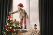 © illustrissima - boy 3-4 years old in red and white stripped pajamas and Santa Claus hat plays on the window sill near the decorated New Year tree with lanterns and gingerbread house. Christmas symbols, holiday card
