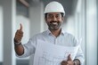 © movinglines.studio - Confident Indian architect with construction plans giving thumbs up in industrial setting.