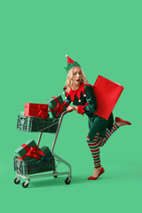 Wall Mural - Beautiful young woman dressed as elf with shopping cart and Christmas gifts on green background