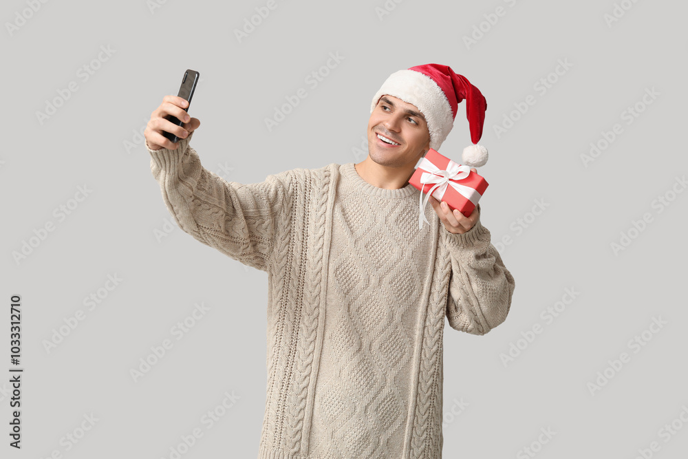 Handsome young man in Santa hat with Christmas gift taking selfie on white background