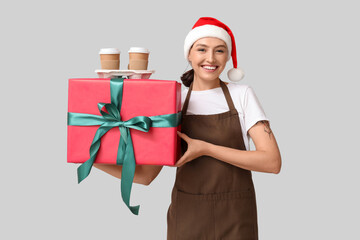 Beautiful female barista in Santa hat with cups of coffee and gift box on grey background