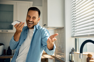 Wall Mural - Cheerful man recording voice message on mobile phone in kitchen.