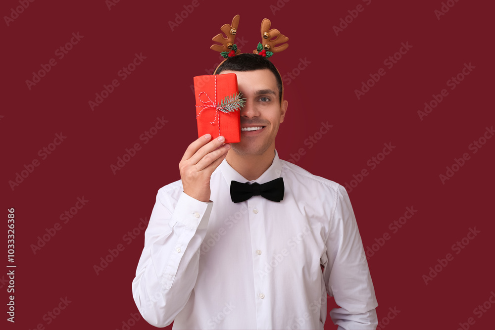 Young man in reindeer horns with Christmas gift box on red background