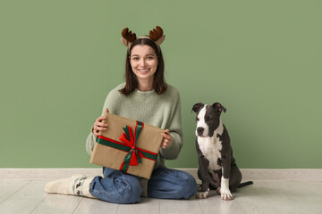 Wall Mural - Beautiful young woman in reindeer horns with Christmas gift and cute Staffordshire Terrier puppy near green wall