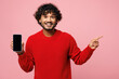 © ViDi Studio - Young Indian man he wear red sweater casual clothes hold in hand use mobile cell phone with blank screen workspace area point finger aside isolated on plain pastel pink background. Lifestyle concept.