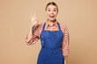 © ViDi Studio - Young happy surprised woman barista bartender barman employee wear blue apron shirt work in coffee shop show okay ok gesture isolated on plain pastel beige background Small business startup concept