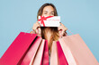 © ViDi Studio - Close up young woman wear hold in hand paper package bags after shopping cover mouth with voucher card wink blink eye isolated on plain light pastel blue background. Black Friday sale buy day concept.