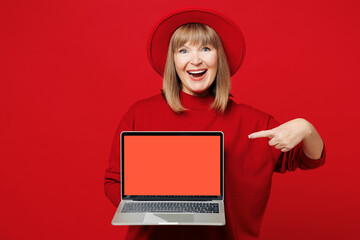 Elderly fun blonde IT woman 50s years old wear sweater hat casual clothes hold use work point finger on laptop pc computer with blank screen area isolated on plain red background. Lifestyle concept.