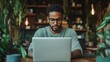 ©  Shomixer - A man with glasses is deeply immersed in his laptop, surrounded by lush green plants and warm lights, creating an atmosphere of concentration and creativity.