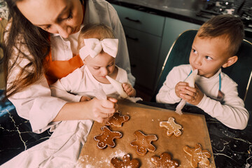 A top view of a black table with gingerbread cookies as a mother and her children decorate them with icing together.