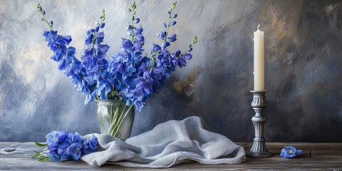 Poster - Tall blue delphinium flowers are arranged in a vase on the table alongside a candlestick and a fabric napkin.