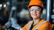 © Pinklife - A young female engineer wears protective glasses and gloves, sporting an orange hard hat and safety vest, standing confidently amidst machinery in a busy warehouse setting.