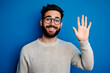 © john - Young handsome man with beard wearing casual sweater and glasses over blue background Waiving saying hello happy and smiling, friendly welcome gesture