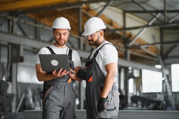 Two Heavy Industry Employees in Hard Hats Discussing Job Assignments at the Factory, Using Laptop Computer