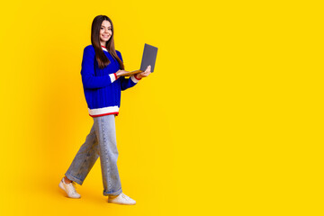 Wall Mural - Full length photo of pretty teen girl hold netbook walking wear trendy knitwear blue outfit isolated on yellow color background