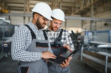 Two international male workers in overalls looking at the laptop against the background of the production of aluminum PVC windows and doors