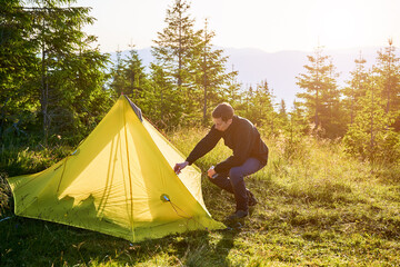 Wall Mural - Camper nearly yellow tent at sunrise in scenic mountain setting. Tent bathed in golden morning light, surrounded by tall evergreens and grassy terrain. Distant mountains and clear sky.