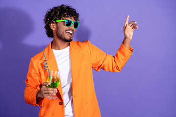 Canvas Print - Photo of attractive funny guy wear orange suit drinking cocktail waving emtpy space isolated violet color background