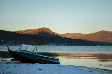 Winter sunset with a pink sky reflecting over Teletskoye Lake in the Altai mountains. Boat on the shore. Serene beauty of a short winter solstice day