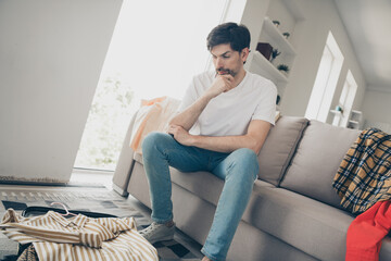 Pensive young man sitting on sofa at home contemplating weekend plans with casual attire choices