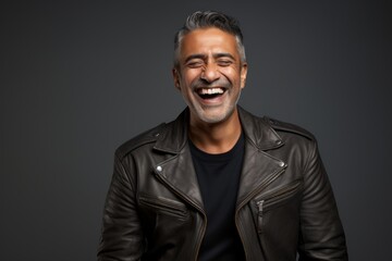 Portrait of a cheerful indian man in his 40s sporting a classic leather jacket while standing against soft gray background