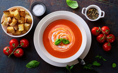 Food photography of a creamy soup of tomato. The tomato soup comes with a swirl of fresh cream in the middle of the table, covered with crispy croutons and a pinch of basil, a plate of fresh tomato le