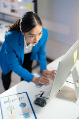 Businesswoman is standing at her desk and working late, typing on her computer with financial charts visible on the desk