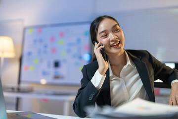 Businesswoman is working late and having a cheerful conversation on her phone. She is sitting at her desk with a laptop, paperwork, and a whiteboard behind her