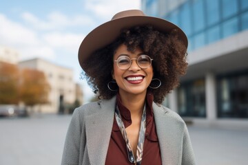 Canvas Print - Portrait of a merry afro-american woman in her 40s donning a classic fedora while standing against modern university campus background