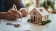 © ksu_ok - person signs a real estate contract with a miniature house and keys on the table, symbolizing property purchase, home ownership, or real estate investment.