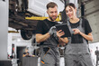 © anatoliycherkas - Young expert female inspects repair checklist with automotive mechanic worker partner, quality suspension technician team at fix garage. Vehicle maintenance service works industry occupation job.