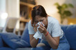 ©  NCST Studio - Young woman is sitting on her couch blowing her nose with a tissue while suffering from a cold or allergies