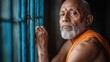 ©  Shomixer - Mature man gazing thoughtfully through window bars, dressed in traditional orange attire, evokes introspection and wisdom, captured in a serene, contemplative moment.