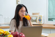 © Ekkasit A Siam - Asian woman drinking orange juice while working on a laptop in kitchen. Fruits arranged on the table nearby, breakfast setting with toast and juice. Multitasking between eating and working in kitchen