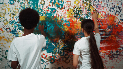 Back view of young diverse children paint colorful stained wall with hand. Attractive highschool student paint the wall with hand print while holding color palette. Creative activity. Edification.