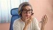 © Sheikhah - Woman with short gray hair applies makeup while holding mirror, white top, blue chair, sitting at white desk, pink wall and plant in background. Scene suggests focus on self-grooming, calm energy.