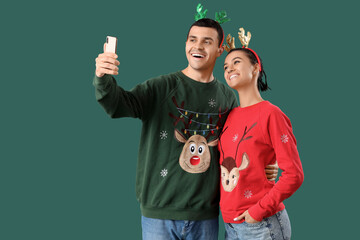 Canvas Print - Young couple in Christmas sweaters and reindeer horns taking selfie on green background