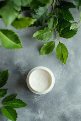Wall Mural - A white jar of moisturizer with creamy texture is placed on a polished grey surface, next to bright green leaves. The simplicity of the grey background highlights the natural elements, promoting a