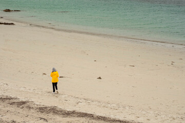 Wall Mural - Teenager in yellow jacket walking on a white stone beach by turquoise ocean. Dog bay, county Galway, Ireland. Travel and tourism. Stunning Irish landscape scene. Enjoy nature theme.