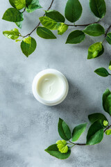 Wall Mural - A white jar of moisturizer with creamy texture is placed on a polished grey surface, next to bright green leaves. The simplicity of the grey background highlights the natural elements, promoting a