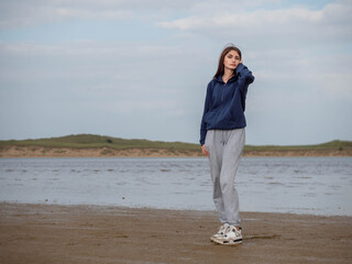 Teenager girl stands on a beach in front ocean. She is wearing a blue hoodie and gray pants. Selective focus. Calm nature scene. Travel and tourism. Model slim body type with long hair. 