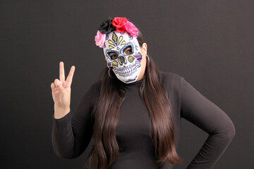 Mexican adult woman with catrina mask shows the sign of love and peace with her hand to celebrate the Day of the Dead, tradition and culture of Mexico